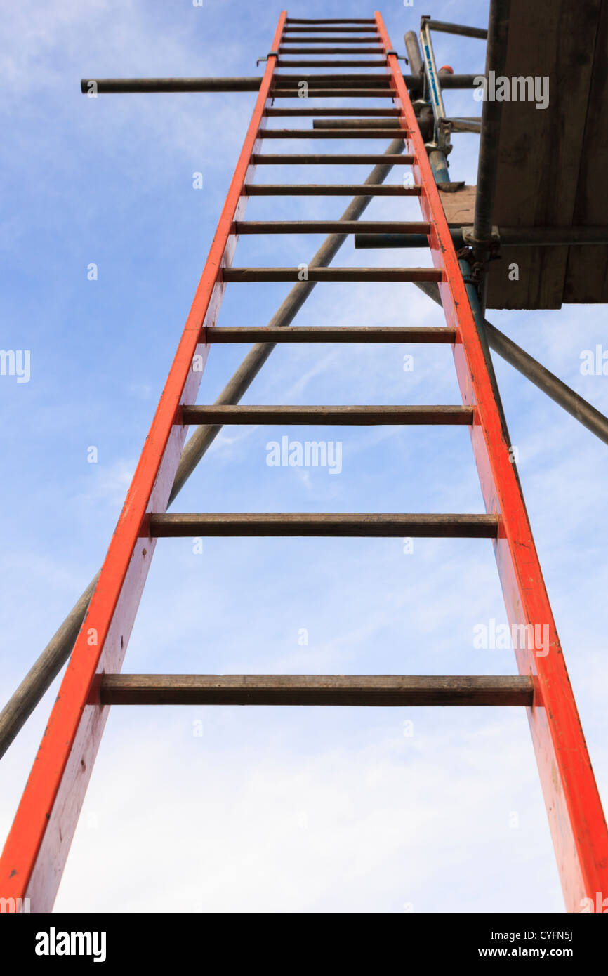 Red step ladder leading up to a scaffolding platform from the bottom rung against blue sky to illustrate ambition and opportunity concept. UK Stock Photo