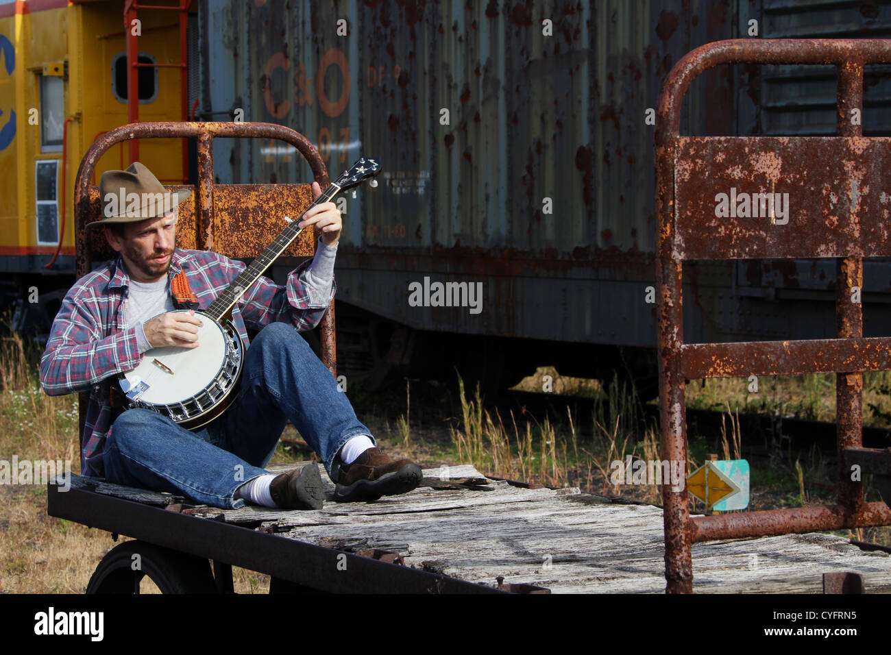 Male model as a hobo playing banjo while sitting on an old railroad baggage cart. Stock Photo