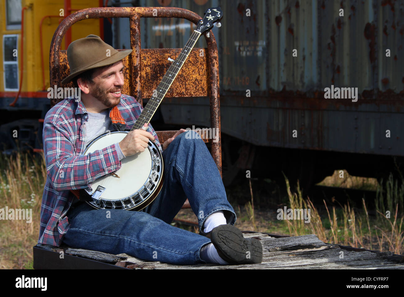 Male model as a hobo playing banjo while sitting on an old railroad baggage cart. Stock Photo