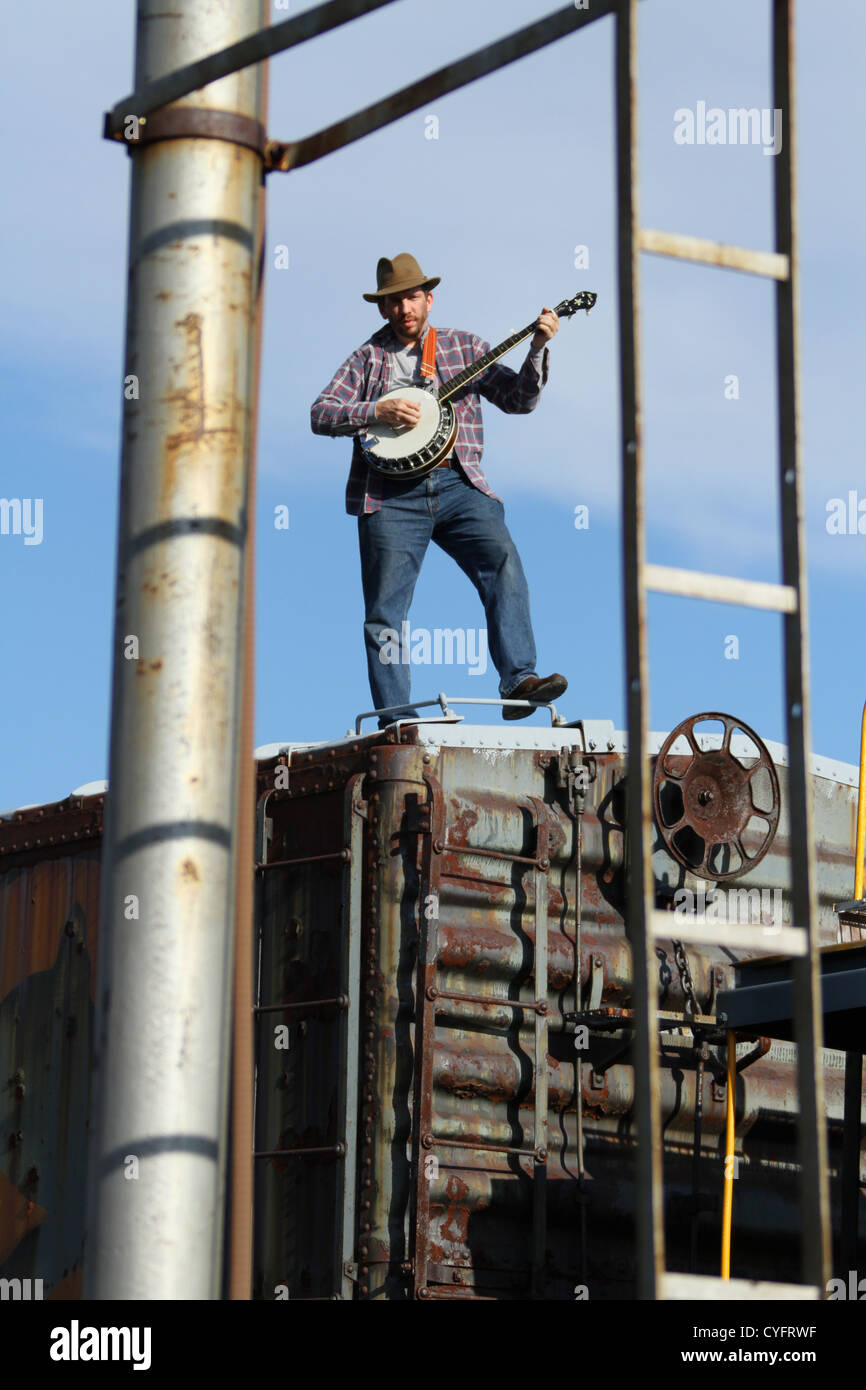 Male model as a hobo playing banjo while standing on a railroad box car. Stock Photo