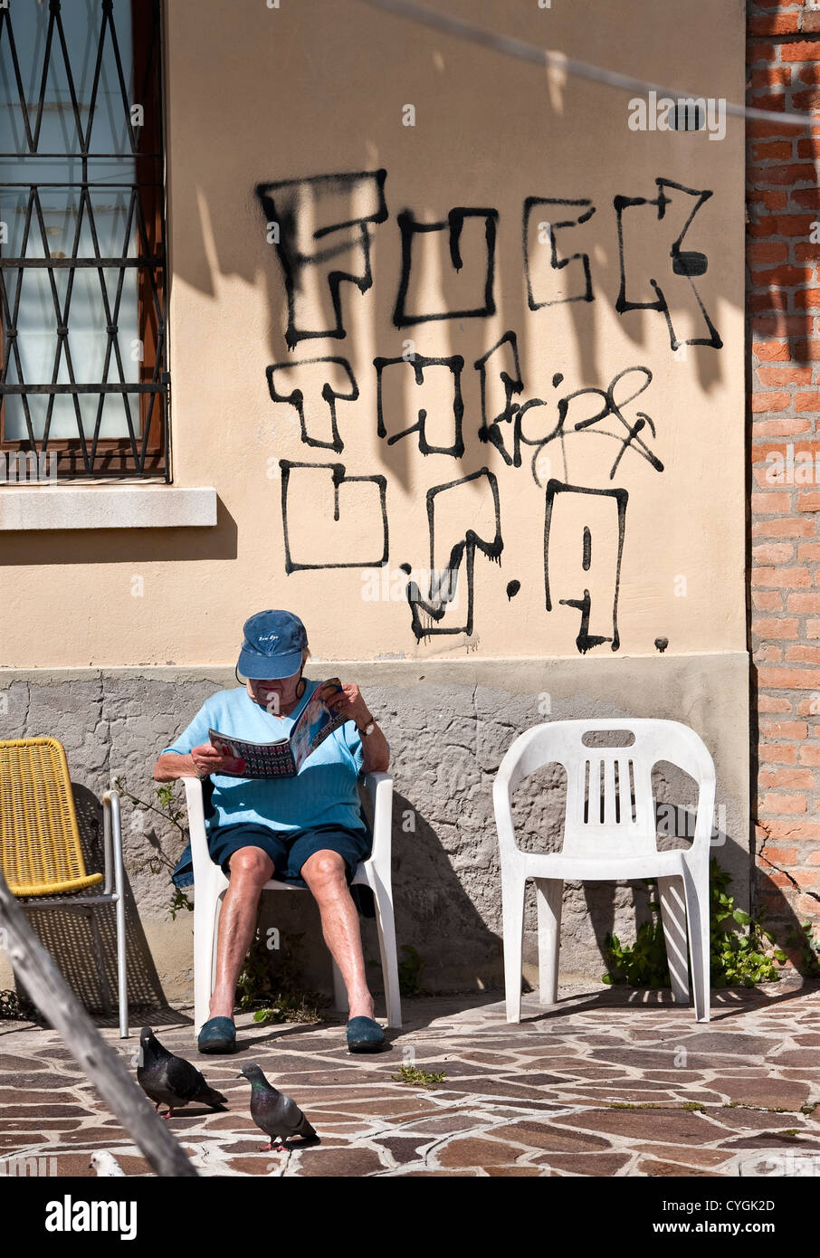 An elderly lady sitting in the sun reading a magazine, beneath some anti-American graffiti in a Communist district of Venice, Italy Stock Photo