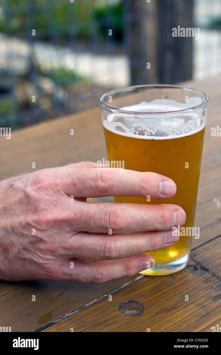Close up of an older man's hand holding a glass of beer on a picnic table. Stock Photo