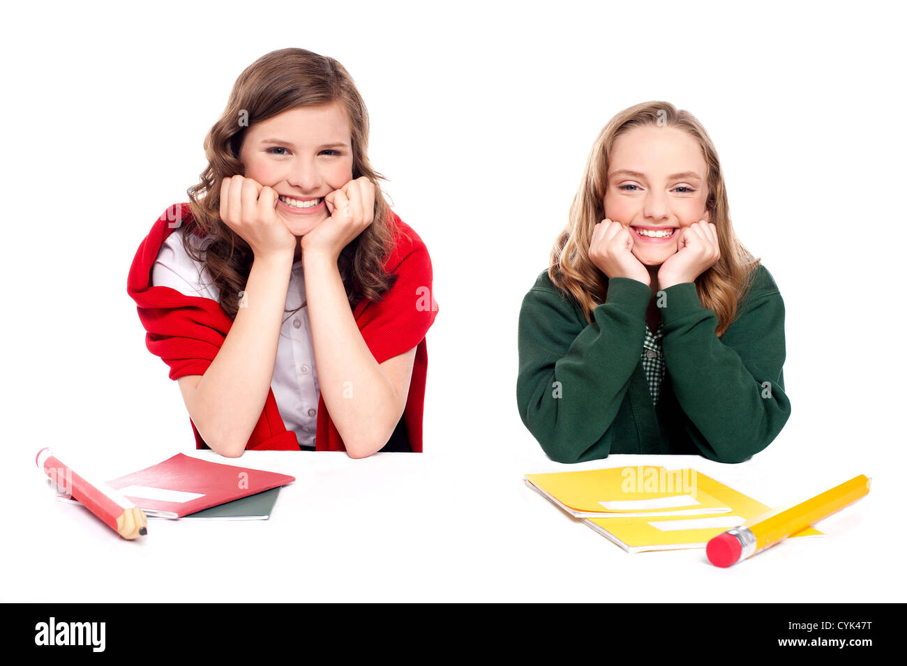 Happy young girls sitting at desk and resting hands on chin isolated on