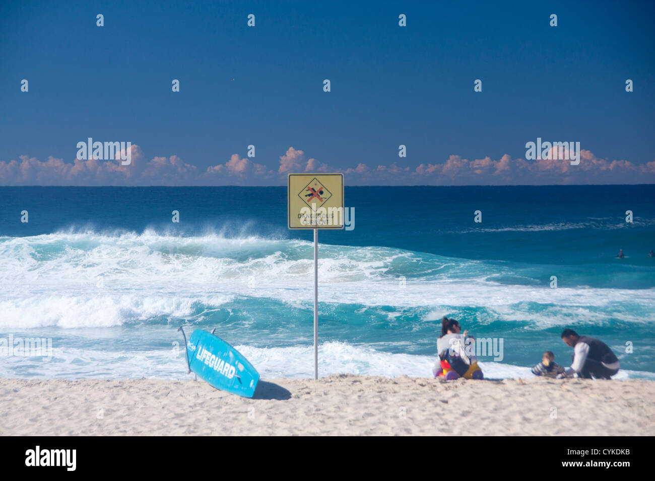 Dangerous Currents sign next to lifeguard surfboard with family playing on beach and stormy sea Bronte Sydney NSW Australia Stock Photo