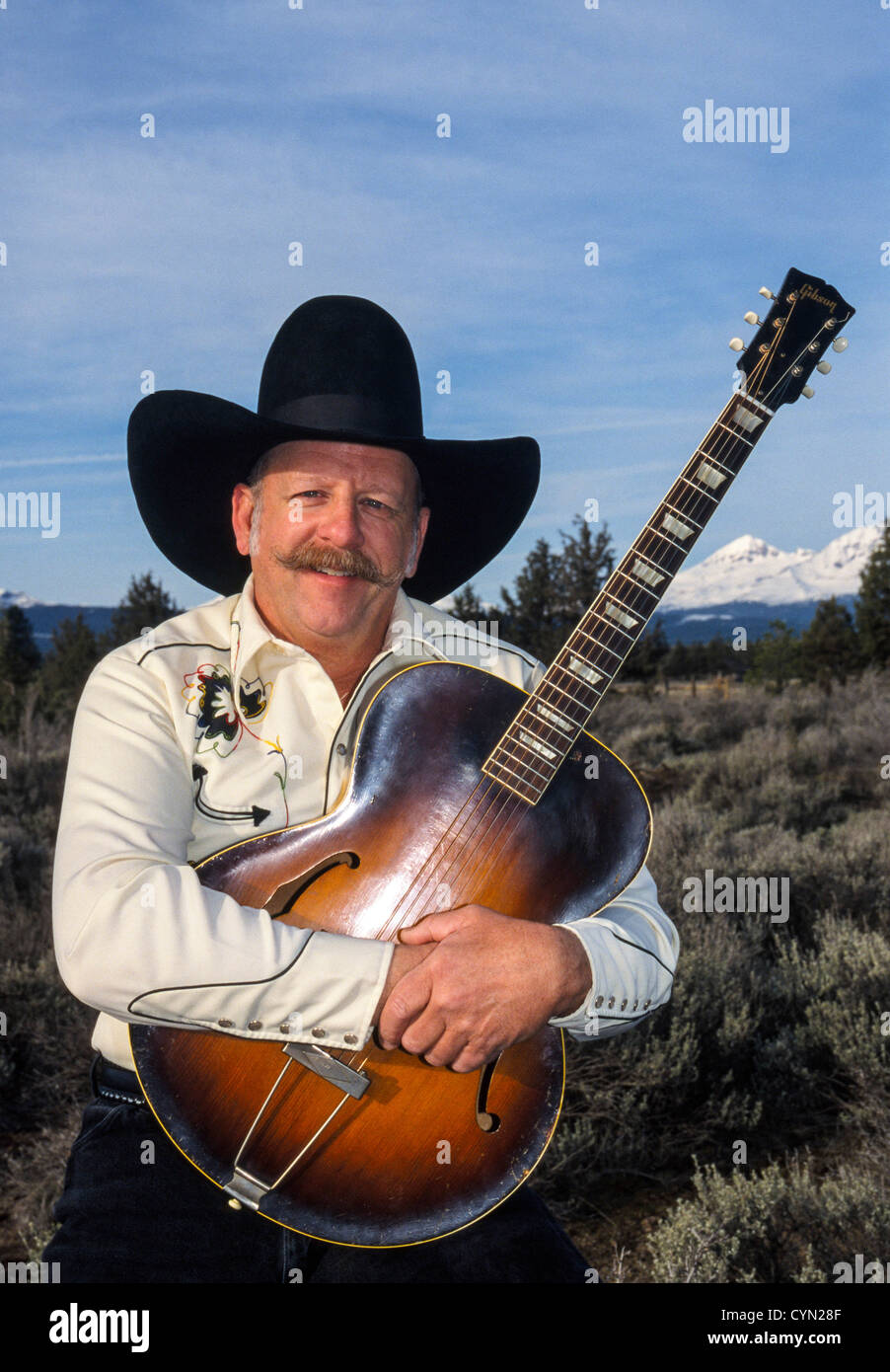 Cowboy and country-western musician John Grant poses with his guitar amid desert sagebrush near the Cascade Mountain Range at Bend, Oregon, USA. Stock Photo