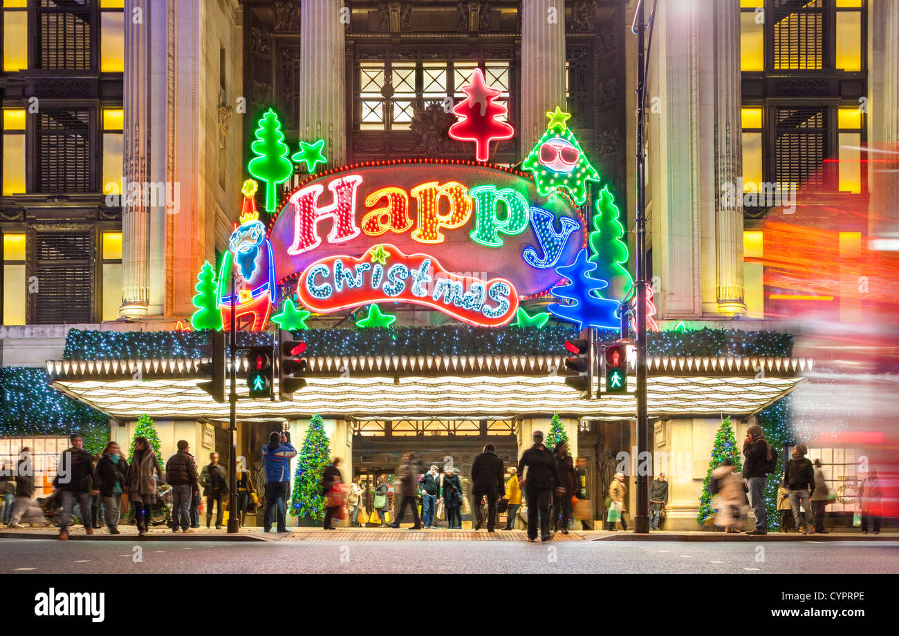 London UK. Christmas shoppers Xmas shopping at Selfridge's store Oxford Street with large neon sign lights Happy Merry Christmas Stock Photo