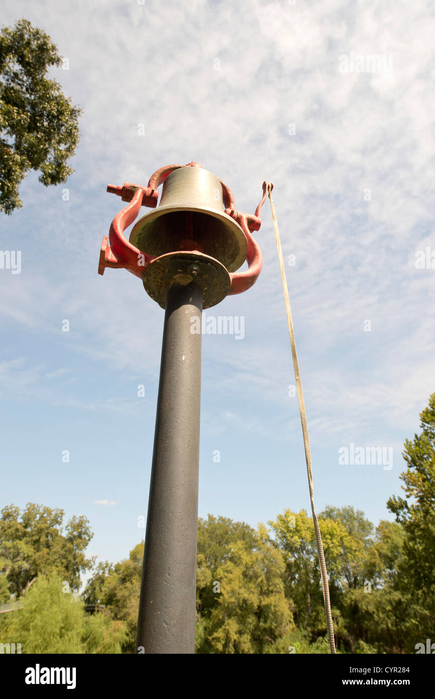 An old metal dinner bell stands on a pole near the San Gabriel River in