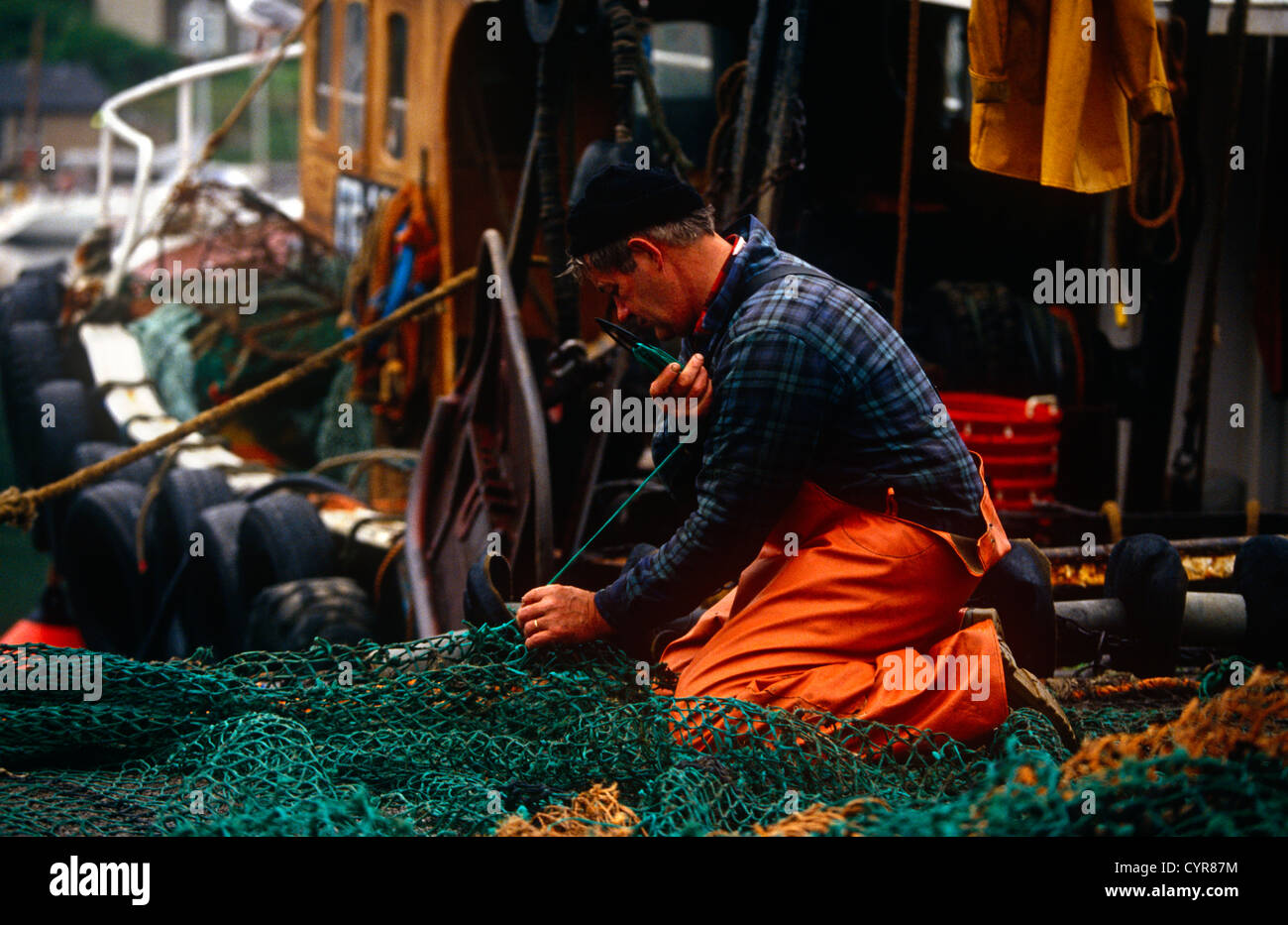 With his boat in the background, a fishermen repairs his nets on the quayside after a night at sea in Tarbert, Mull of Kintyre. Stock Photo