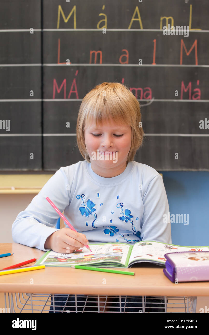 Fresh first year schoolgirl student is sitting at the desk, working with textbook and smiling. Stock Photo