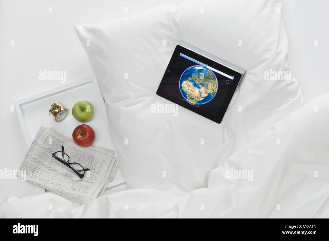 Digital tablet, alarm clock, apple, spectacles with newspaper on bed