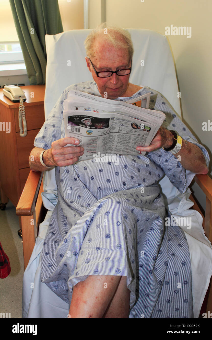 a senior elderly man in a gown sitting in a chair reading a newspaper in his hospital room in a hospital or clinic Stock Photo