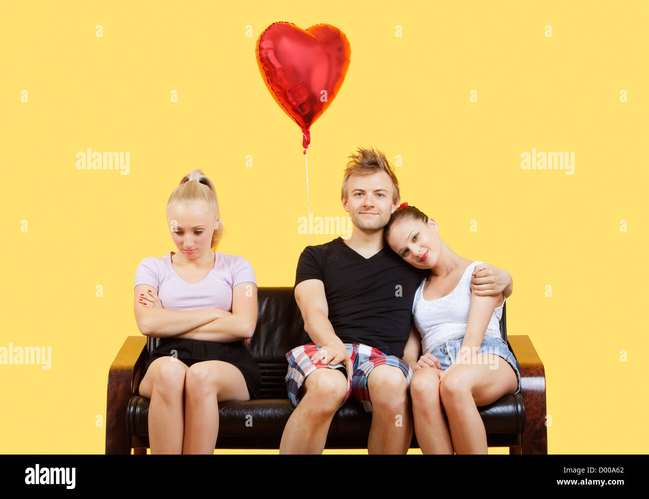 Portrait of young couple sitting on sofa with female friend feeling left out over yellow background Stock Photo