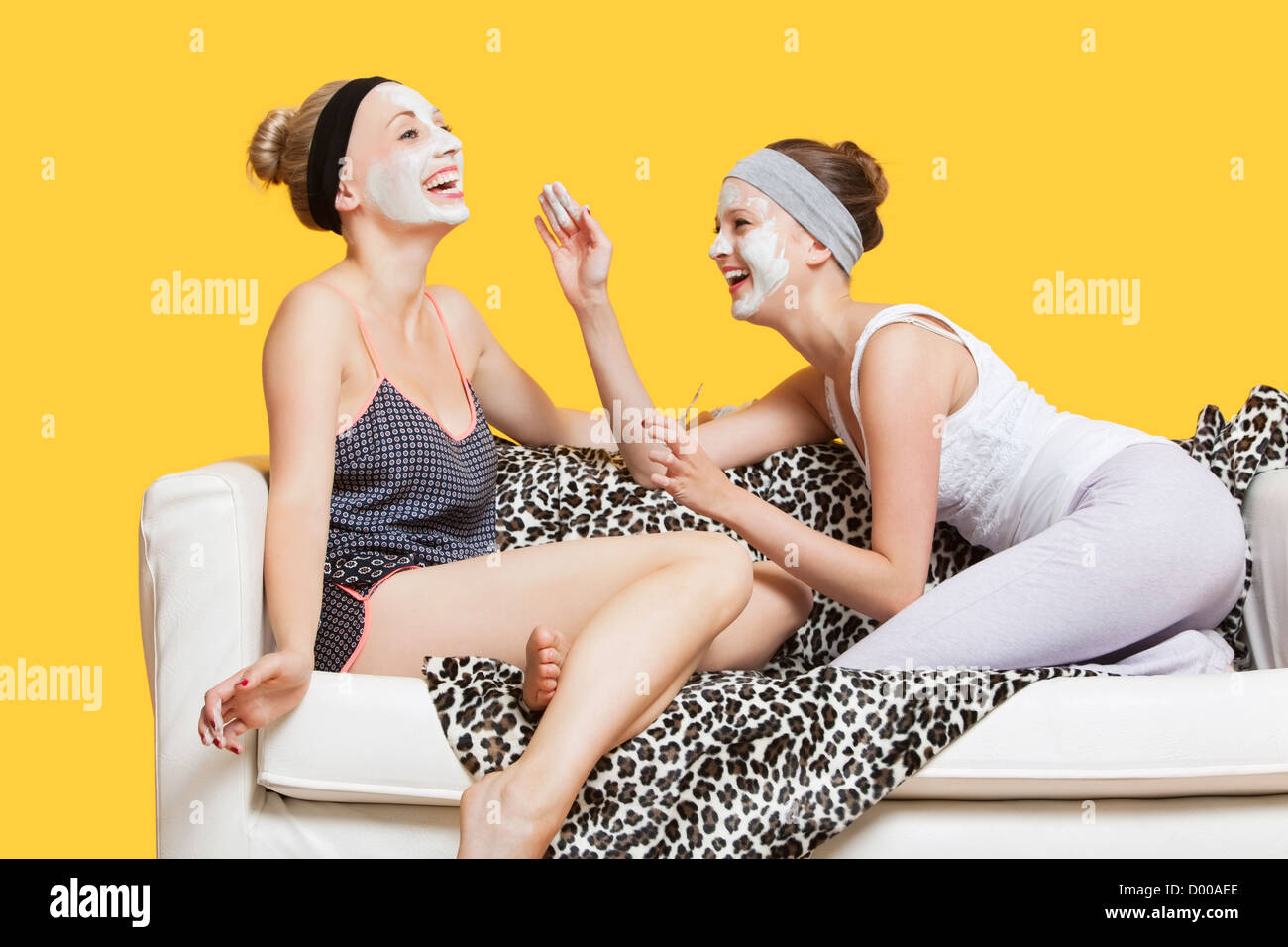 Two happy young women applying face pack while sitting on sofa over yellow background Stock Photo