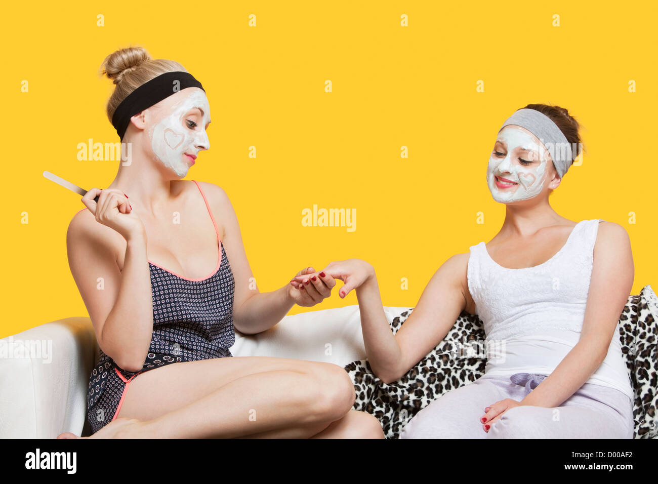 Young woman with face pack filing friend's nails while sitting on sofa over yellow background Stock Photo