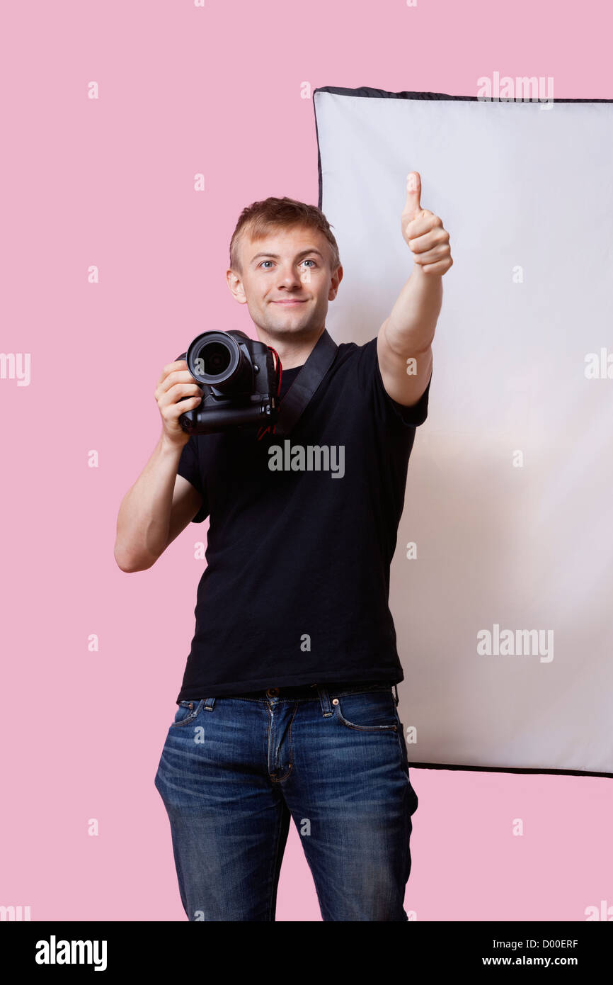Happy photographer holding camera with thumbs up gesture over pink background Stock Photo