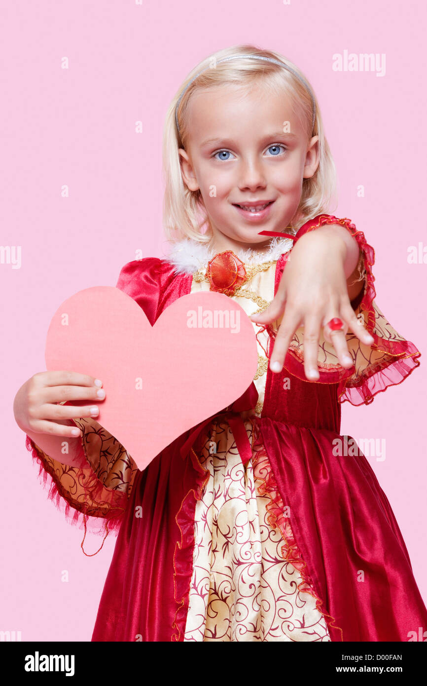 Portrait of a happy young girl dressed in princess costume holding paper heart as she displays ring over pink background Stock Photo