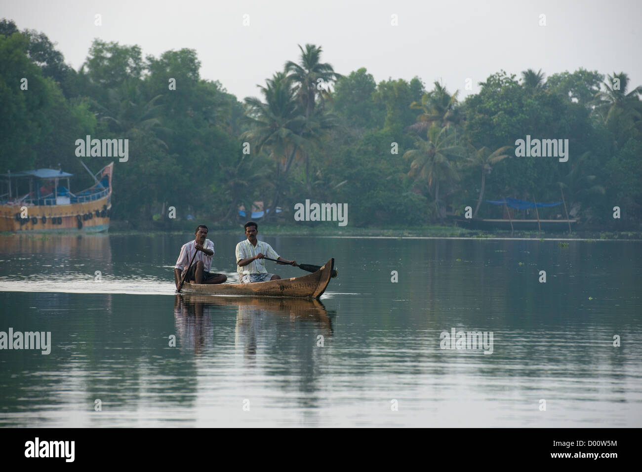 Two men rowing a small canoe past large coloured fishing boats on the West Coast Canal (National Waterway No 3), Kanjippadom, near Alappuzha (Alleppey), Kerala, India Stock Photo
