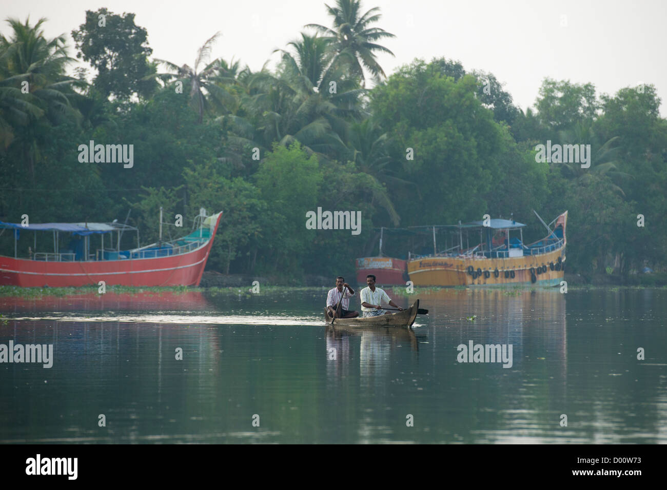 Two men rowing a small canoe past large coloured fishing boats on the West Coast Canal (National Waterway No 3), Kanjippadom, near Alappuzha (Alleppey), Kerala, India Stock Photo