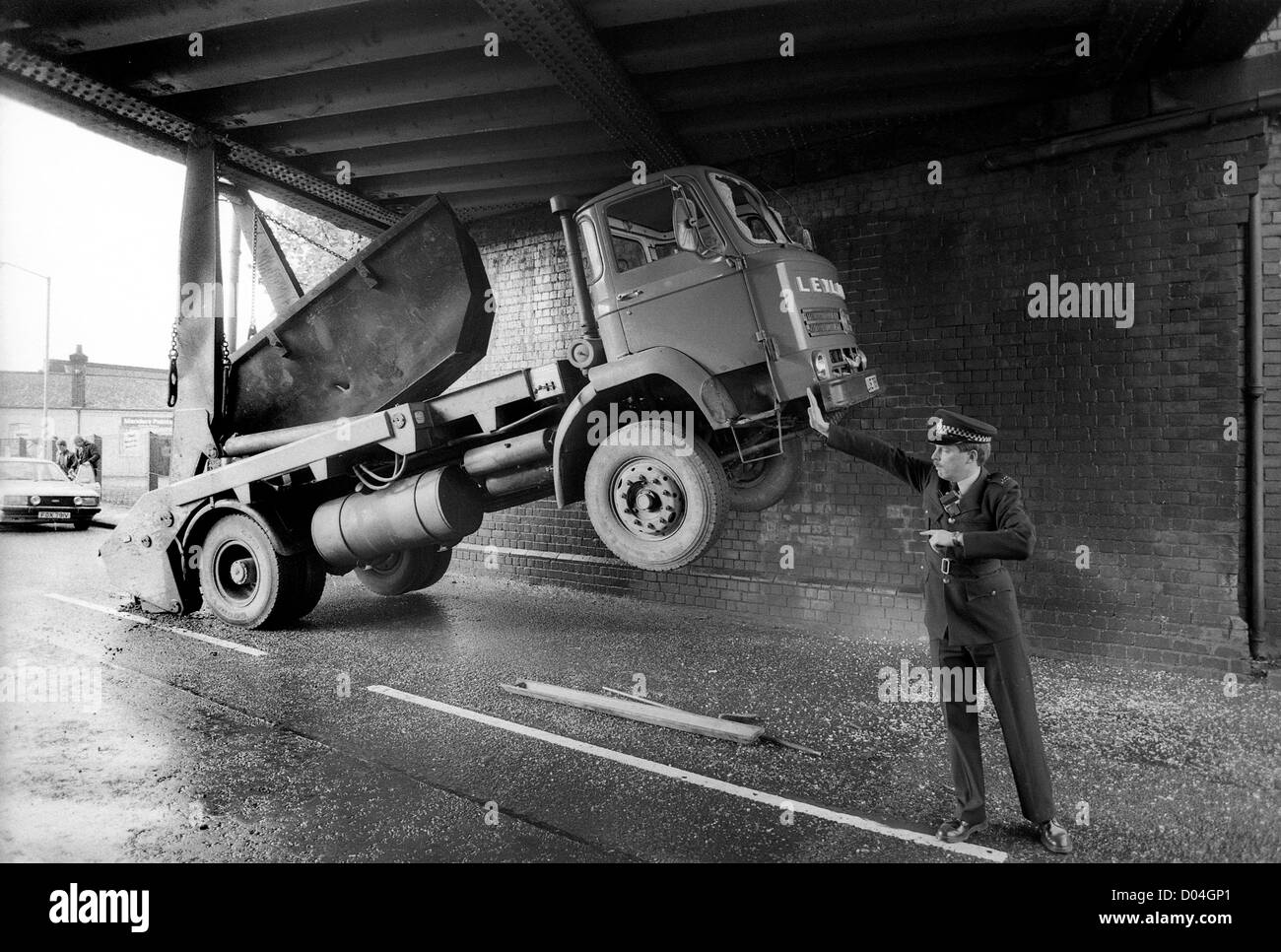 A skip lorry wedged underneath a bridge appears to be held up by the hand of a policeman as he controls traffic 1985. Britain 1980s road blocked accident police officer traffic collision truck stuck low bridge height Stock Photo