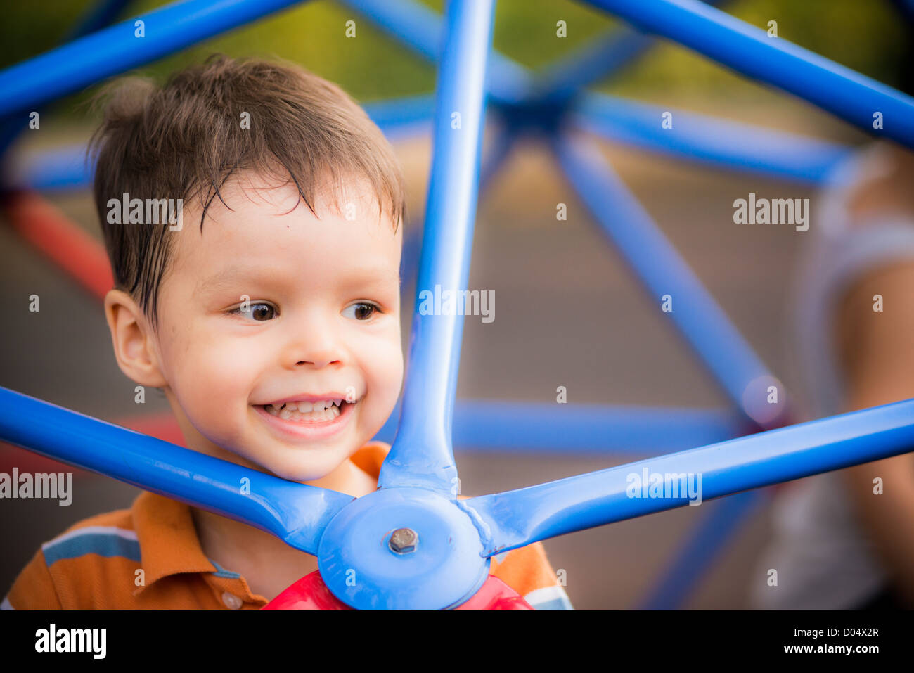 A cheerful 2 year old boy plays on blue monkey bars in outdoor playground Stock Photo