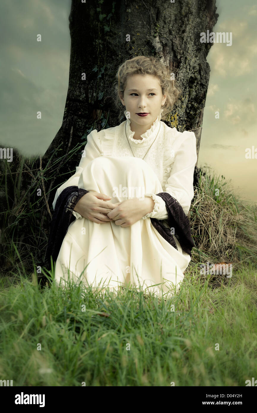 a girl in a victorian dress is sitting under a tree Stock Photo