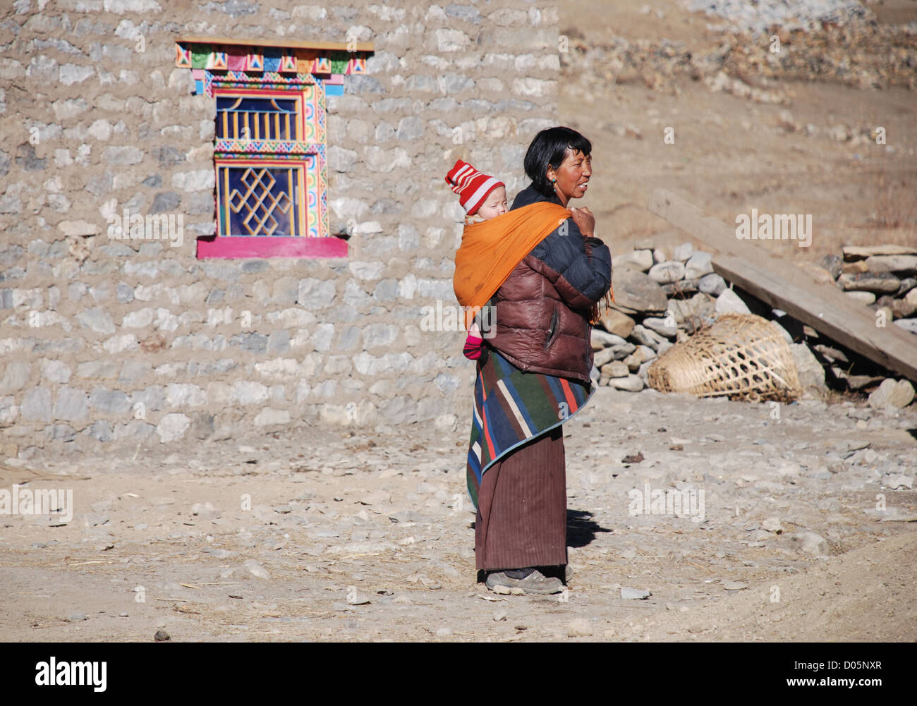 A woman wearing traditional Tibetan dress and  carrying a small baby stands outside a house in the dolpo region of Nepal Stock Photo