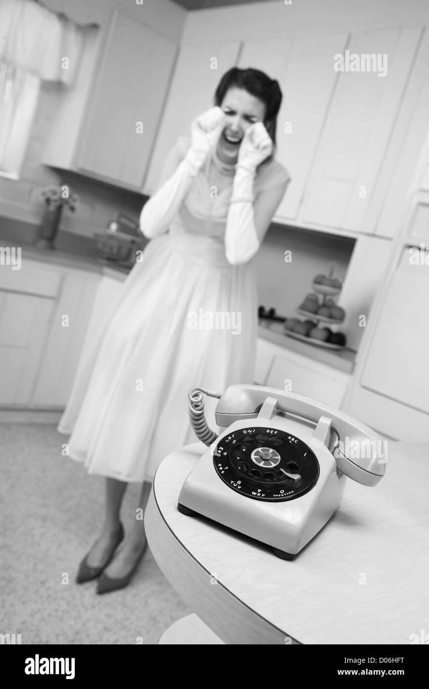 Young Caucasian woman weeps near a phone in a retro-style kitchen scene Stock Photo