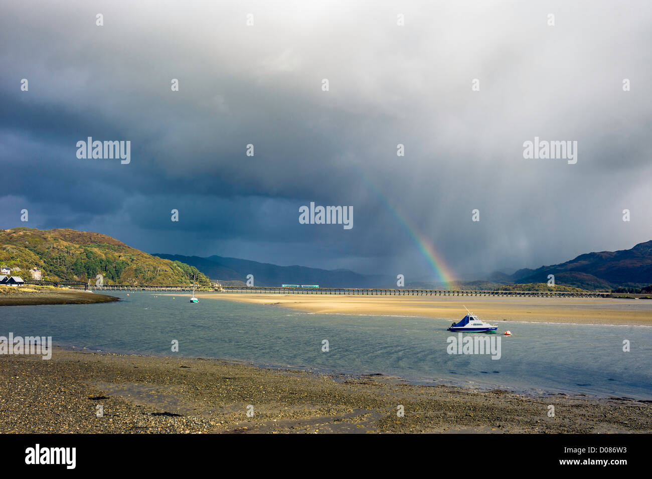local commuter train passes over Barmouth estuary bridge, under a rainbow, storm over mountains, boats moored in foreground sun Stock Photo