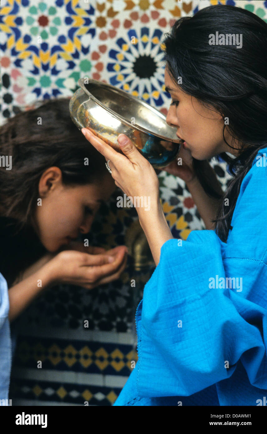 YOUNG MOROCCAN WOMEN IN DJELLABAS DRINKING WATER OUT TASSA AT FOUNTAIN IN HAMMAN TRADITIONAL BODY CARE MOROCCO AFRICA Stock Photo