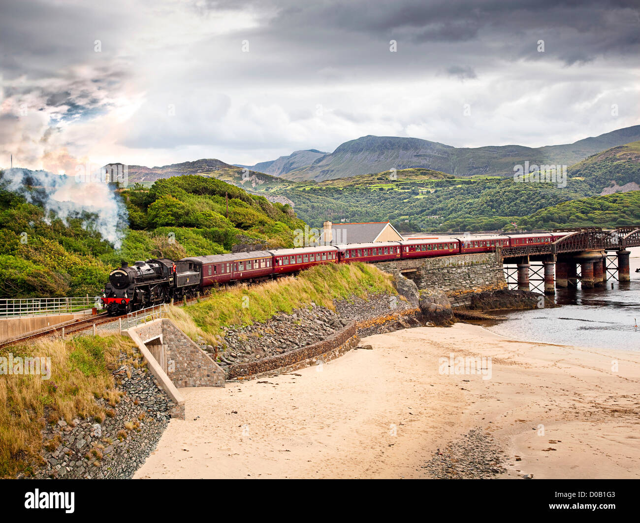 Steam train the Cambrian crosses Barmouth rail bridge over the Mawddach estuary with mountains in the background, near beach. Stock Photo