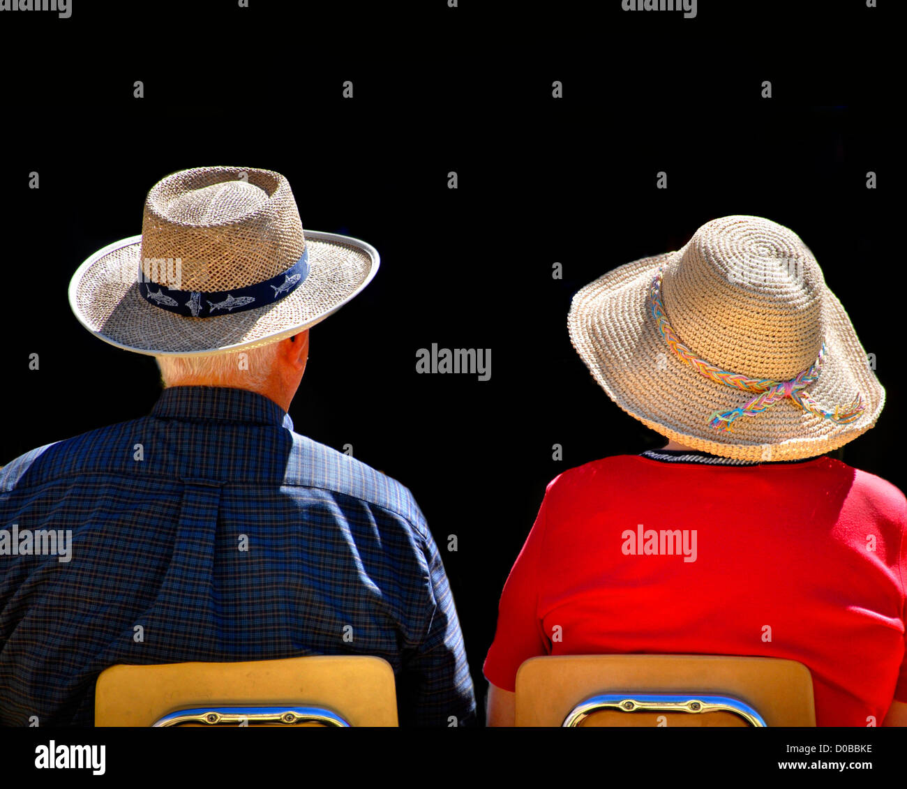 Old couple sitting in chairs wearing straw hats with black background Stock Photo