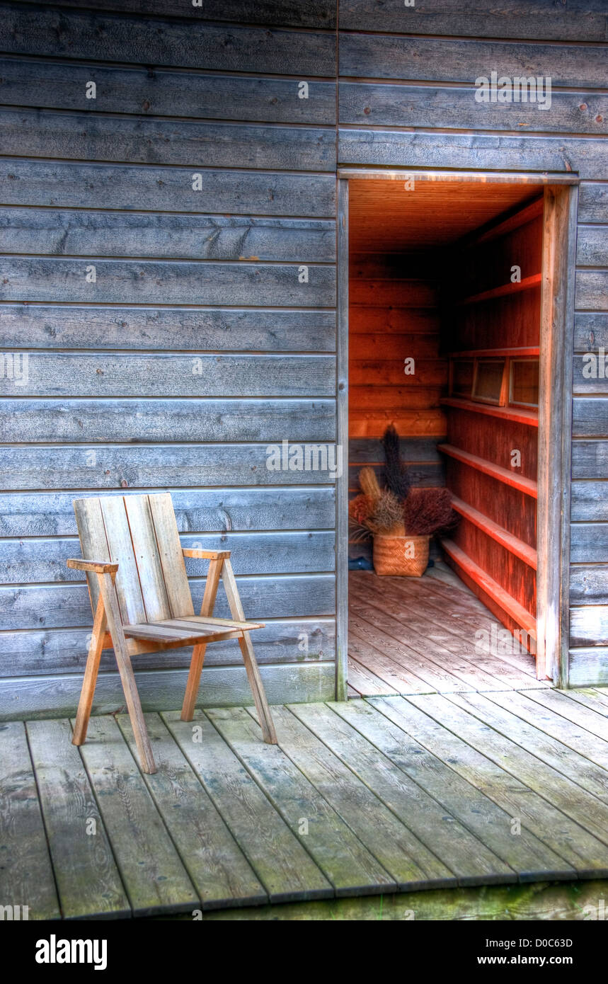 a wooden chair on the wooden patio of a wooden building of Scandanavian design with a doorway opening to a warm interior Stock Photo