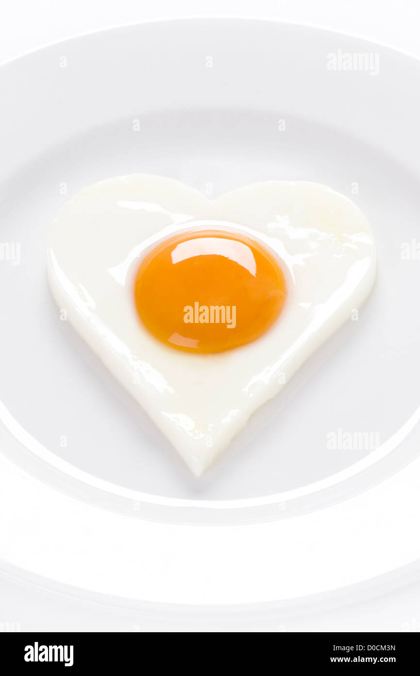 fried egg or poached egg in the shape of a heart on a white circular shaped plate. Egg presented soft 'sunny side up' 'eggs up' Stock Photo