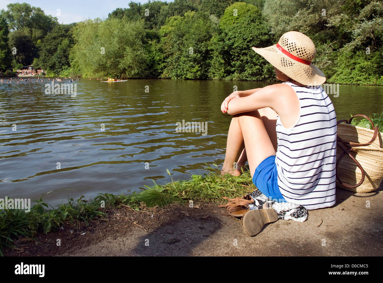 A young lady sunbathes in a wide hat on the banks of the Mixed Pond on Hampstead Heath, north London, on a summers day. Stock Photo