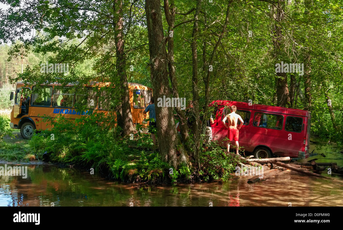 Yellow school bus tows red minibus from broken wooden bridge. Stock Photo