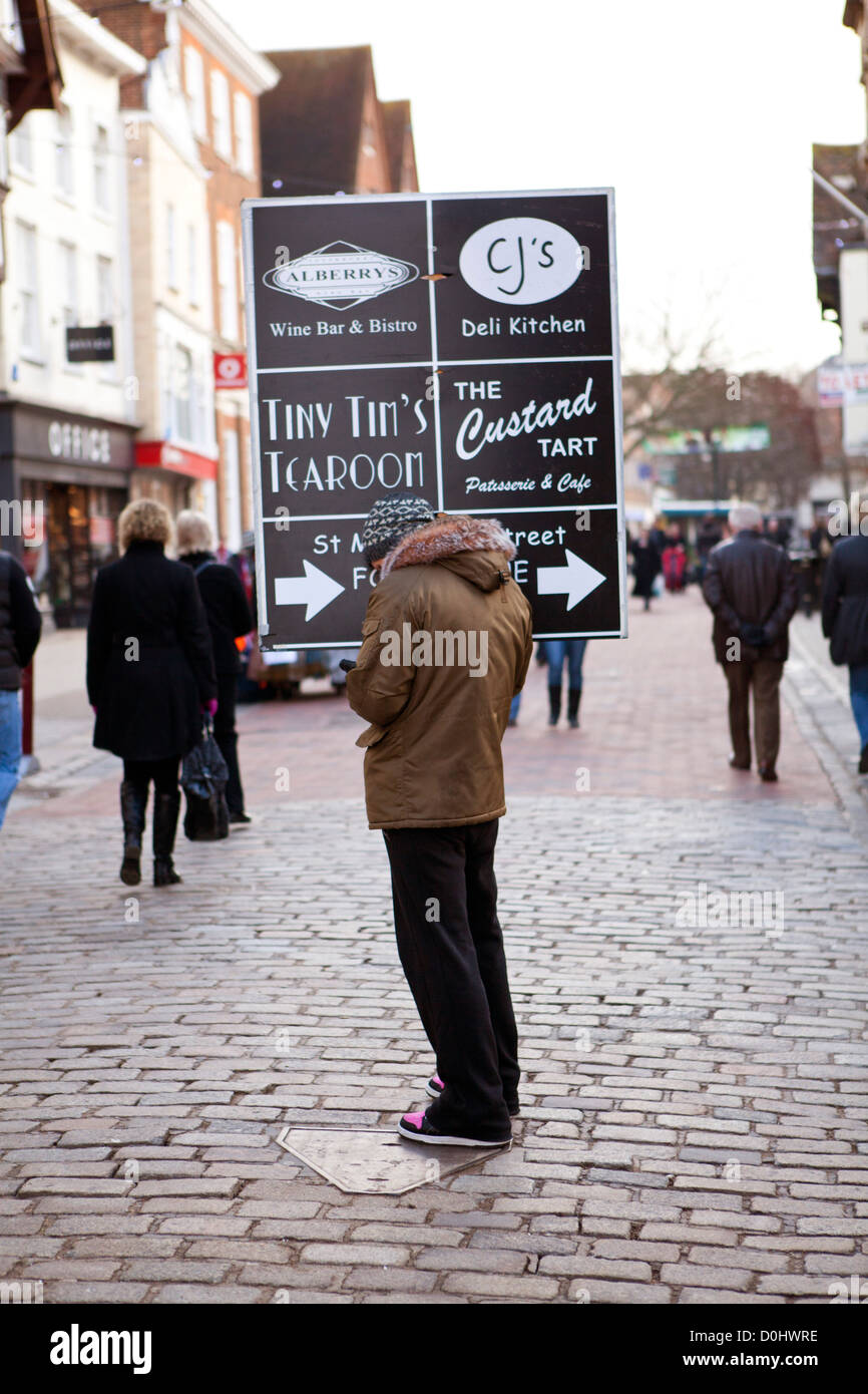 A man stood in the high street holding a placard with directions for local shops. Stock Photo