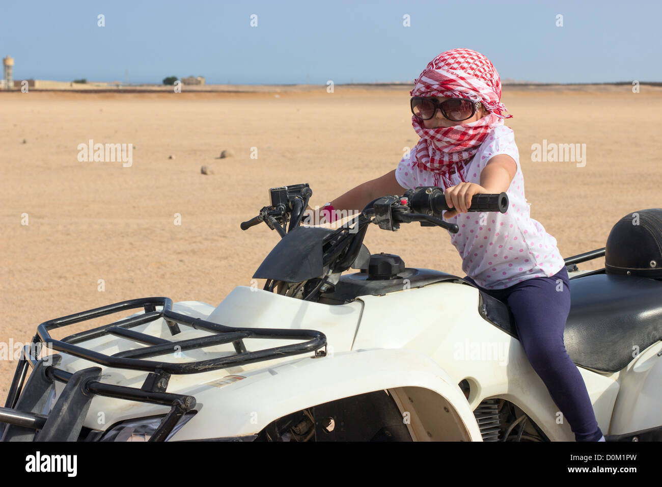 Little Caucasian girl in head kerchief and sunglasses on quad bike in desert Stock Photo