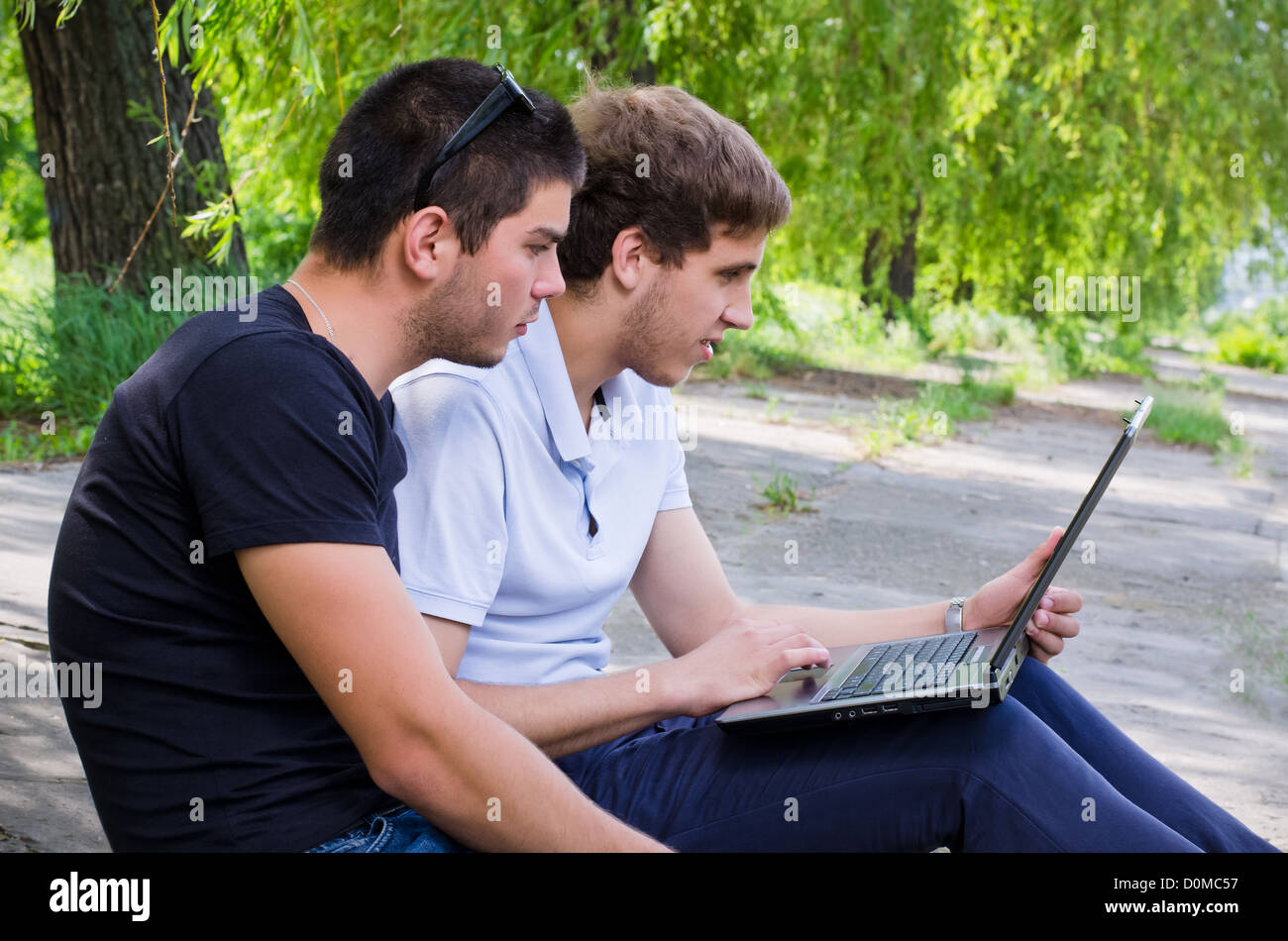 Two young men sitting on the ground outdoors under trees using a laptop together Stock Photo