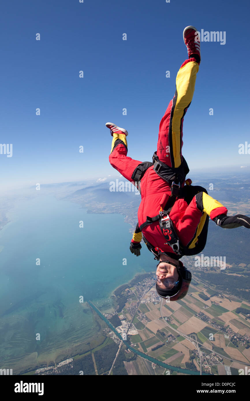 Skydiver girl is smiling in a head low position in the blue sky over a beautiful lake area. Stock Photo