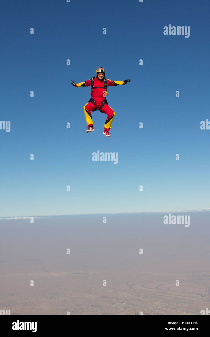 Skydiver girl in a stand-up position is falling free through the blue sky over a dry desert area with a smile on her face. Stock Photo