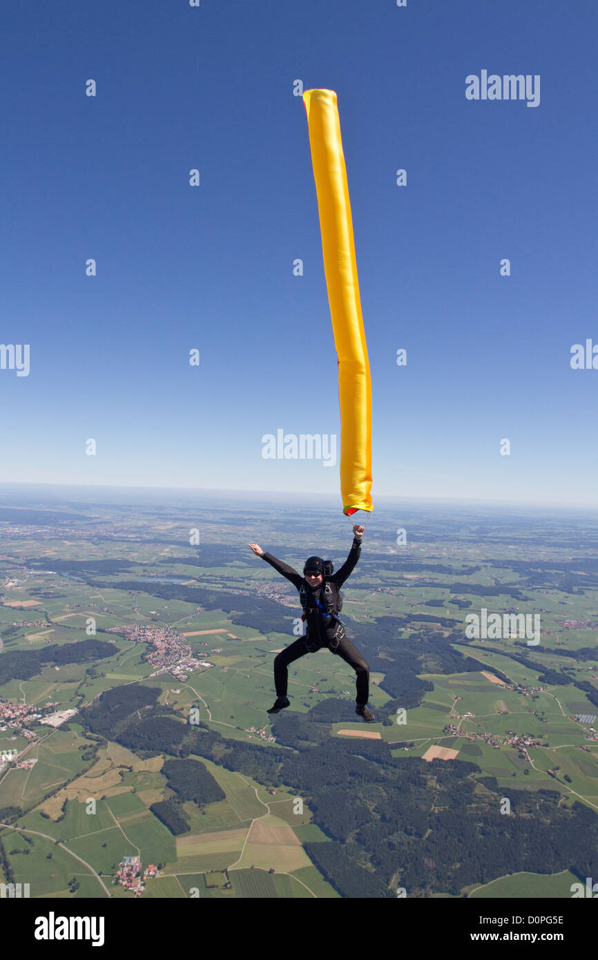 Skydiver is holding a colorful air tube in his hand and falling free through the blue sky over green fields and having fun. Stock Photo