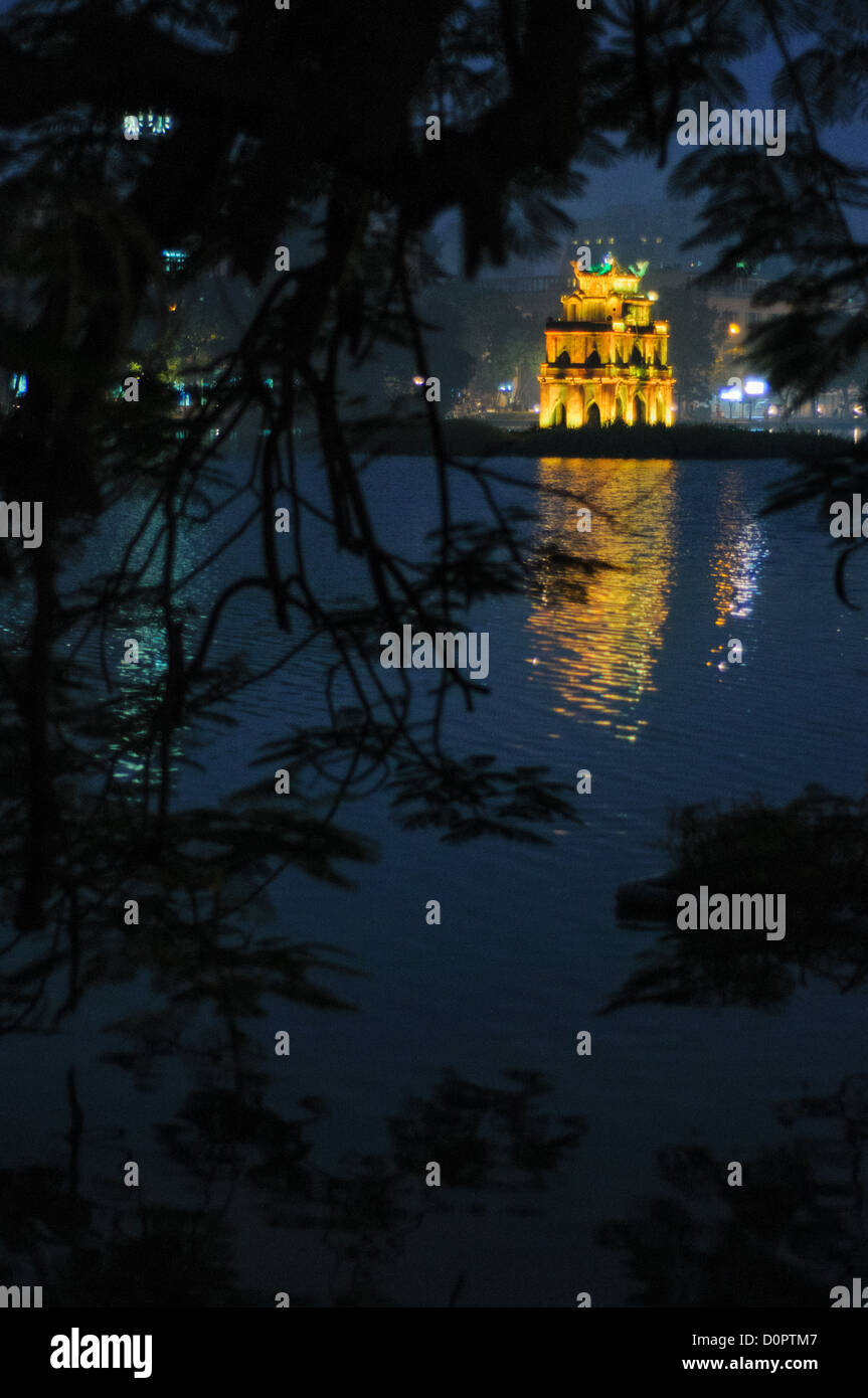 HANOI, Vietnam - Turtle Tower (also known as Tortoise Tower on a small island in Hoan Kiem Lake in the historical center of Hanoi, Vietnam. The lights of the tower are on and reflected on the water, with the image framed by the branches of a tree on the shore. Stock Photo