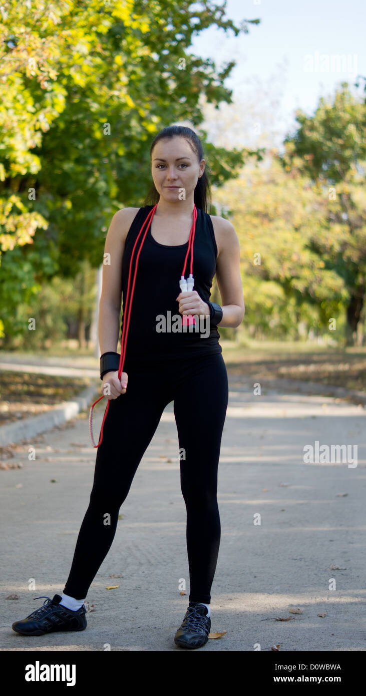 Fit attractive woman in sportswear standing with a red skipping rope draped around her neck in a park Stock Photo