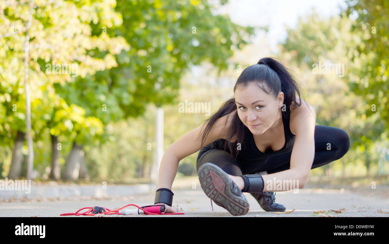 Low angle view of a young attractive woman athlete stretching with her leg extended close to the road surface and a skipping rope in her hand. Stock Photo