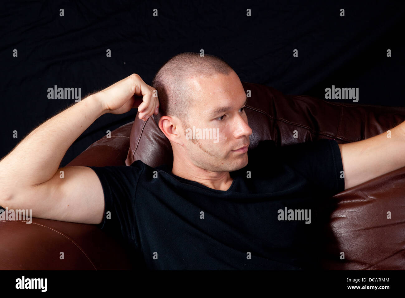 Man with close cut hair sitting on the corner of a couch and looking to the right with his head resting on his hand Stock Photo