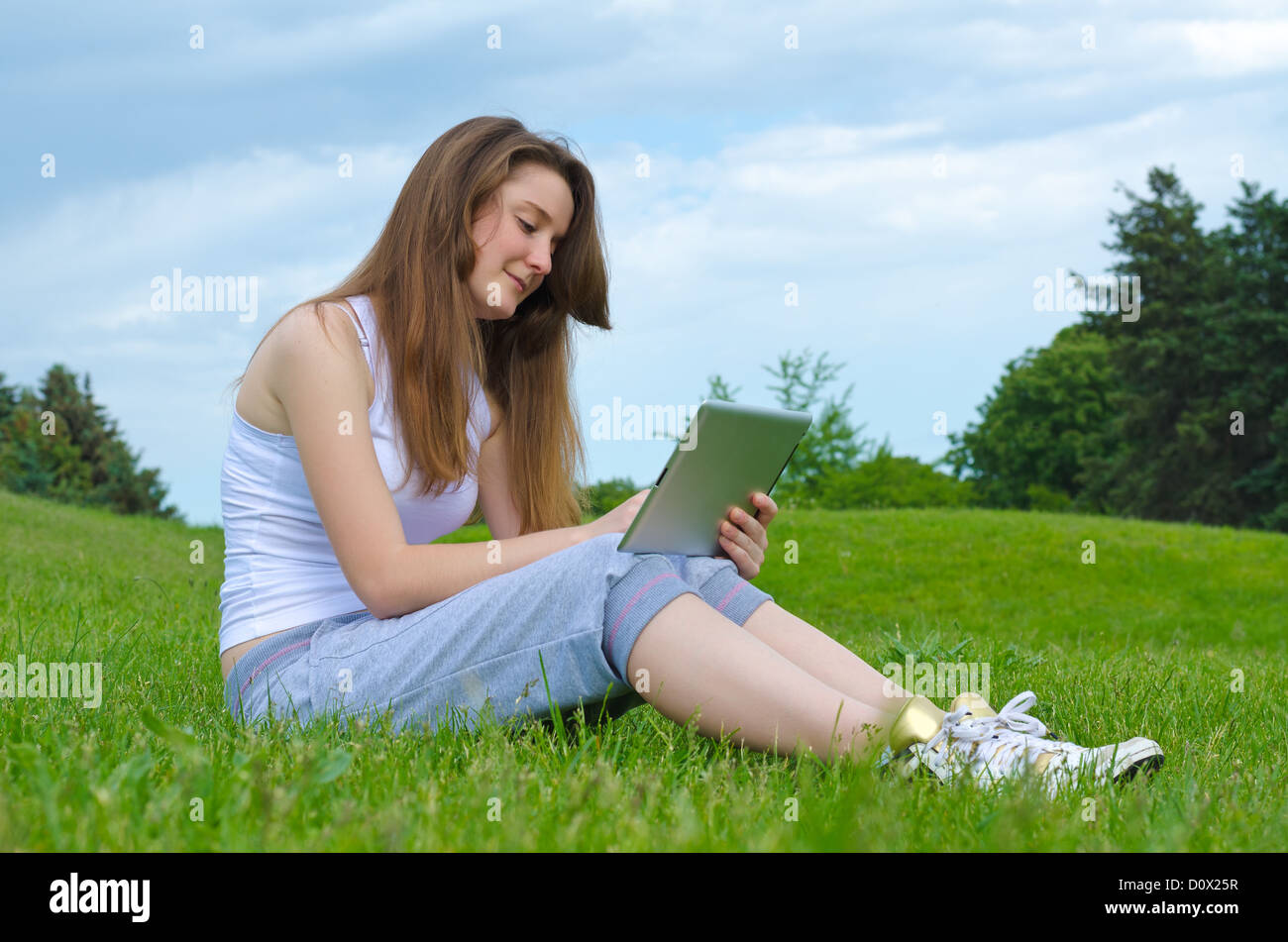 Smiling woman sitting on grass in a lush green park using a touchscreen tablet Stock Photo