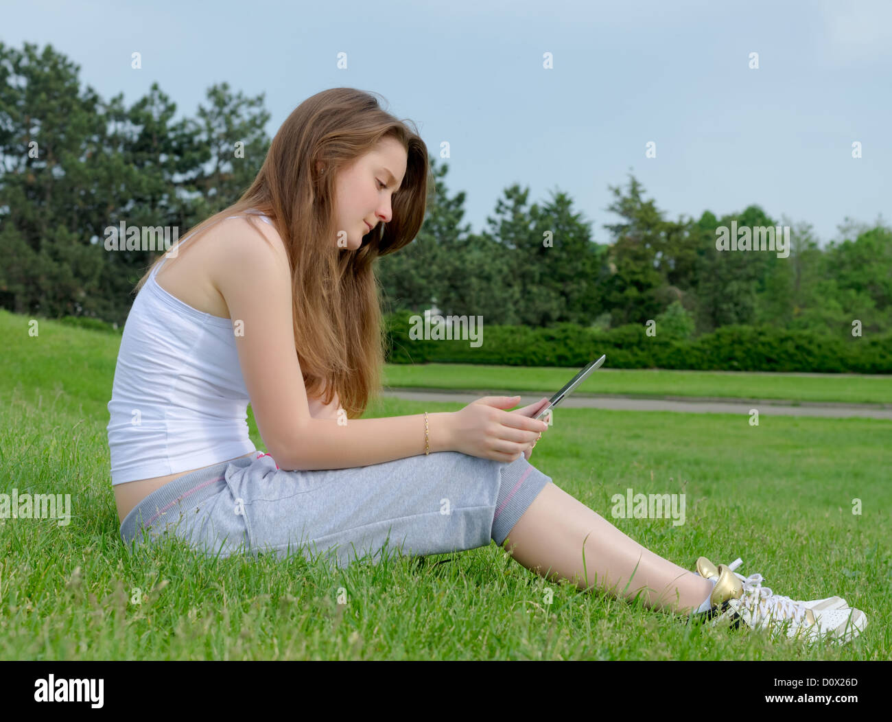 Side view of a young woman sitting in green grass working on a touchscreen tablet in the park Stock Photo