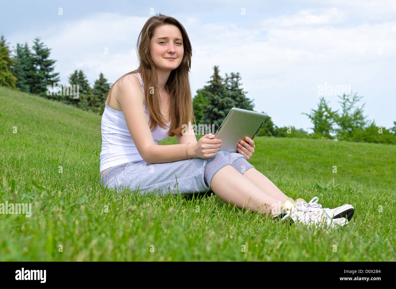 Happy smiling young woman using a touchpad tablet sitting outdoors in the sunshine on a green lawn Stock Photo