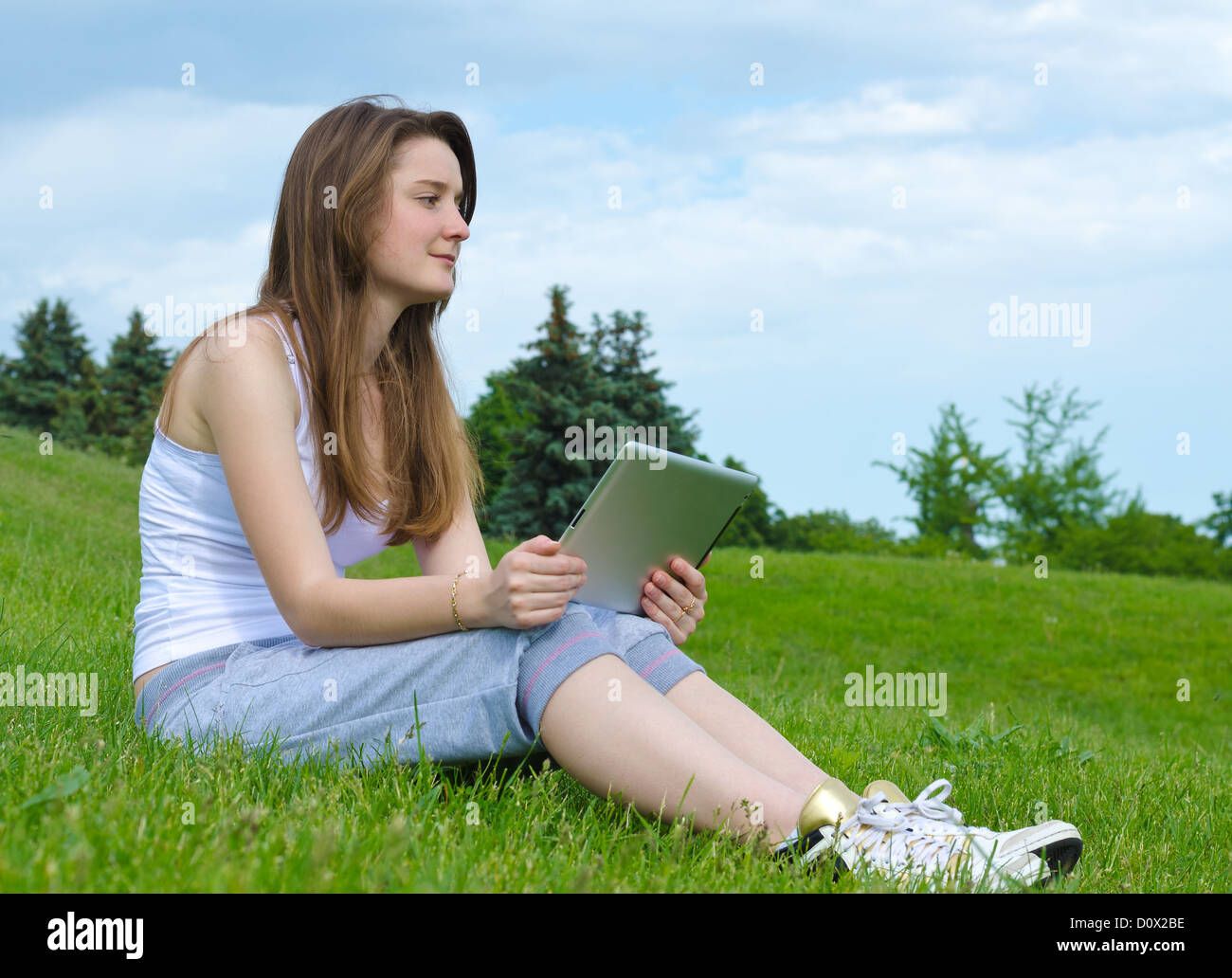Woman sitting on sloping green grass thinking and staring into the distance with a tablet notebook in her hands Stock Photo