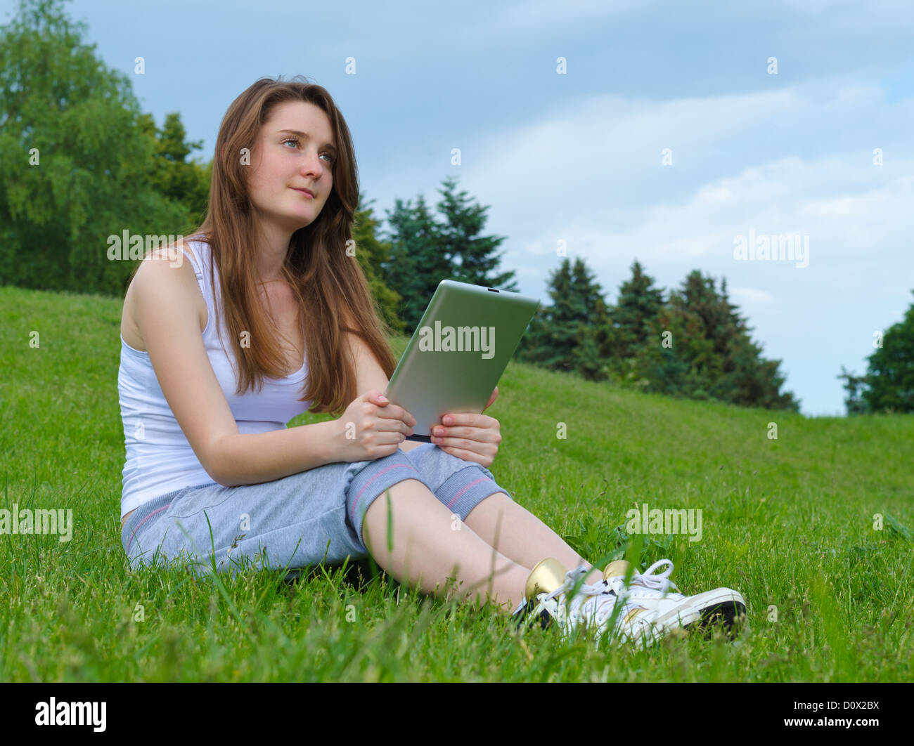 Pensive young woman sitting on green grass staring into the distance while holding a tablet on her knees Stock Photo